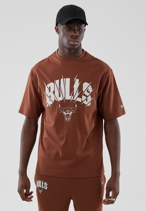Άνδρας με καφέ μπλουζάκι Chicago Bulls και αντίστοιχα σορτς, με μαύρο καπέλο με το λογότυπο των Bulls, στέκεται μπροστά σε απλό γκρι φόντο.