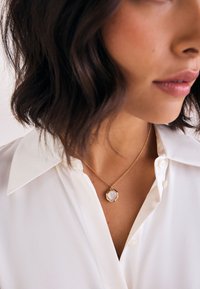 Woman with shoulder-length dark hair wearing a white collared shirt and a gold necklace with an octagonal pink pendant.