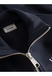 Marineblå zip-up jakke med teksturert krage, sølvfarget glidelås, og en diskret etikett. Stoffet ser glatt og matt ut.
