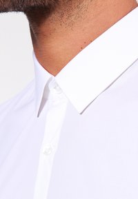 Camisa blanca de cuello clásico con una textura suave, presenta un solo botón en el cuello y no tiene patrones ni acentos visibles.