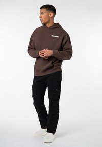 Braune Kapuzenjacke mit "WORLDWIDE"-Logo, kombiniert mit schwarzen Cargo-Hosen mit mehreren Taschen und weißen Sneakers. Glatte Stofftextur.