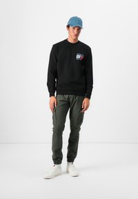 Zwart sweatshirt met een bedrukking op de borst, gecombineerd met olijfgroene cargobroeken met zijzakken, en witte sneakers.