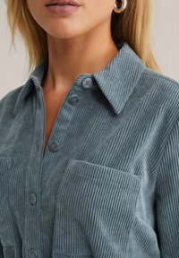 Chemise en velours côtelé bleu ardoise, avec un col et quatre boutons, ainsi que deux poches plaquées. La texture est côtelée et douce.