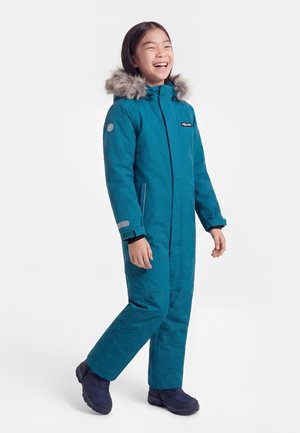 Copil zâmbind, purtând un costum de iarnă turcoaz cu glugă căptușită cu blană și cizme negre, stând în picioare și făcând un pas ușor înainte.