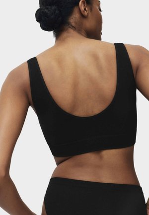 Donna con chignon intrecciato che indossa un reggiseno sportivo nero e slip neri, vista da dietro su uno sfondo chiaro e uniforme.