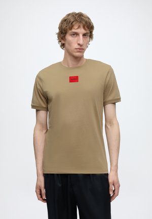 Jeune homme aux cheveux bouclés châtain clair portant une chemise beige à manches courtes avec un patch rouge rectangulaire « HUGO » sur la poitrine.