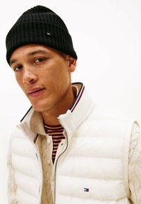 Gilet in bianco con berretto nero a coste. Il gilet presenta una chiusura con zip, un'accenno blu navy e rosso al colletto, indossato sopra un maglione in morbido punto intarsiato color crema.