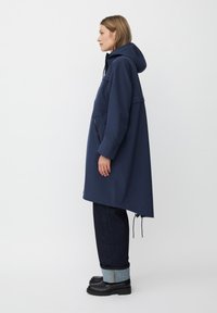 Cappotto blu navy con cappuccio, caratterizzato da una vestibilità ampia, due tasche laterali e un orlo posteriore più lungo. Realizzato in un tessuto morbido e texturizzato.
