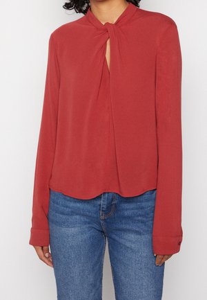 Vrouw met een roestkleurige blouse met lange mouwen en een geknoopt sleutelgat aan de hals, en een blauwe spijkerbroek.