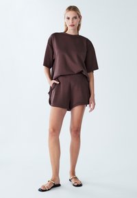 Bruine oversized gebreide t-shirt met korte mouwen, gecombineerd met bijpassende bruine shorts. Model draagt platte sandalen met gouden accenten.