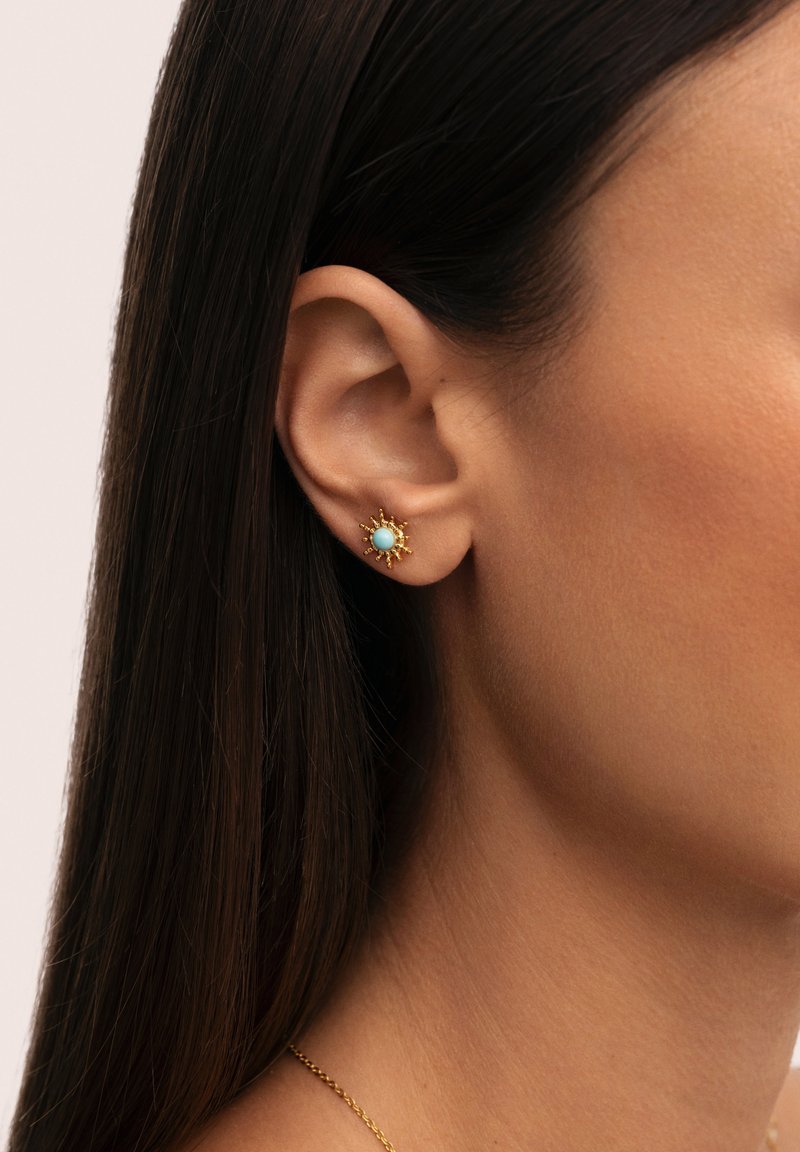Acercamiento al oído de una mujer que lleva un pendiente de oro con un centro de turquesa, con el cabello lacio y oscuro recogido detrás de la oreja.
