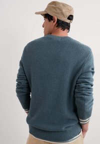 Pull bleu en maille texturée avec col rond, poignets et ourlet côtelés ; présente des rayures blanches et grises à la base et aux manches.