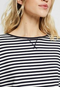 Maglia a righe con strisce orizzontali blu navy e bianche, presenta uno scollo a U e cuciture nere in rilievo sul colletto e sulla patta.