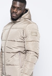 Giacca a vento beige imbottita con cappuccio, caratterizzata da cuciture orizzontali, una tasca con zip e un'etichetta di marca sul braccio sinistro. Texture liscia.