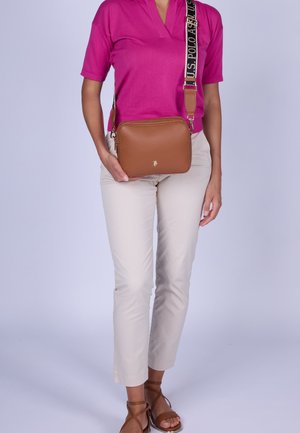 Person wearing bright pink short-sleeve top, light beige pants, brown strappy sandals, and holding brown crossbody bag with black U.S. Polo strap.