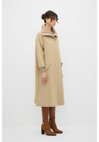 Beige mid-lange jas met een hoge kraag, grote knopen aan de voorkant en zijzakken. Gladde stof met een relaxte pasvorm. Gepaard met bruine kniehoge laarzen.