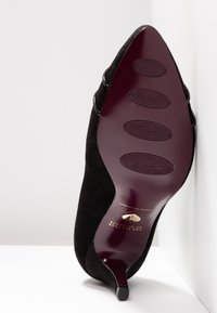 Sorte ruskind højhælede sko med en smal spids tå, der har en tekstureret mønstret sål og let udsmykning på siderne.