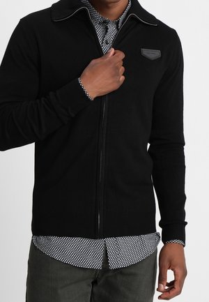 Homme portant un pull noir à fermeture éclair par-dessus une chemise à motif noir et blanc, ajustant le pull près de la poitrine.