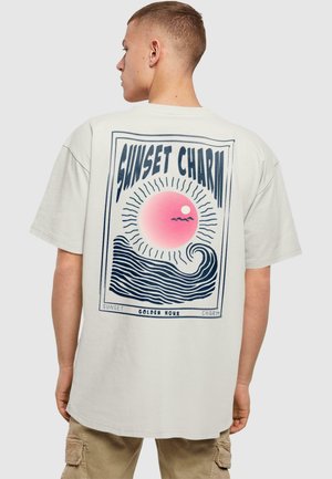 White cotton t-shirt featuring a large graphic print of a sunset, waves, and the text “SUNSET CHARM” in blue and pink colors on the back.