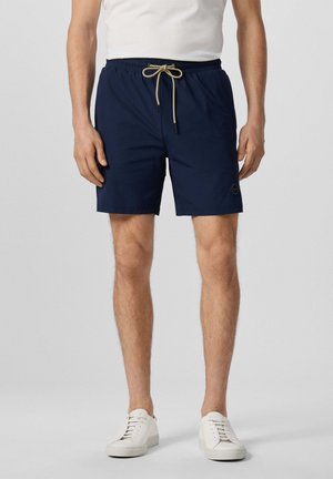 Man draagt marineblauwe shorts met trekkoord en witte sneakers en staat tegen een effen achtergrond.
