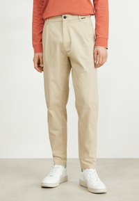 Chino beige con una vestibilità su misura, dotati di zip e chiusura a bottone, abbinati a sneakers bianche e una camicia a maniche lunghe color ruggine.