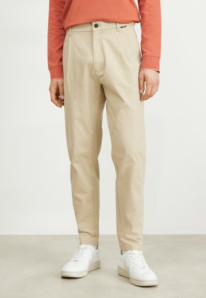 Chino beige con una vestibilità su misura, dotati di zip e chiusura a bottone, abbinati a sneakers bianche e una camicia a maniche lunghe color ruggine.