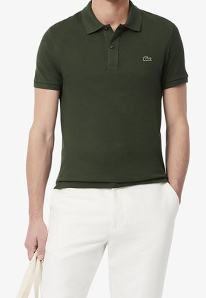 Homme portant un polo vert foncé à manches courtes avec un petit logo crocodile et un pantalon blanc, tenant un sac cabas beige.