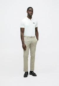Weißes Poloshirt mit Logo, kombiniert mit beigen Chinos und schwarzen Schuhen. Das Model steht aufrecht mit den Händen in den Taschen vor einem einfarbigen Hintergrund.