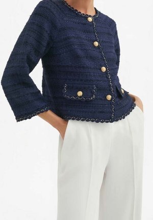 Kobieta w granatowej, teksturowanej kurtce z złotymi guzikami i białych spodniach, z rękami w kieszeniach, na białym tle.