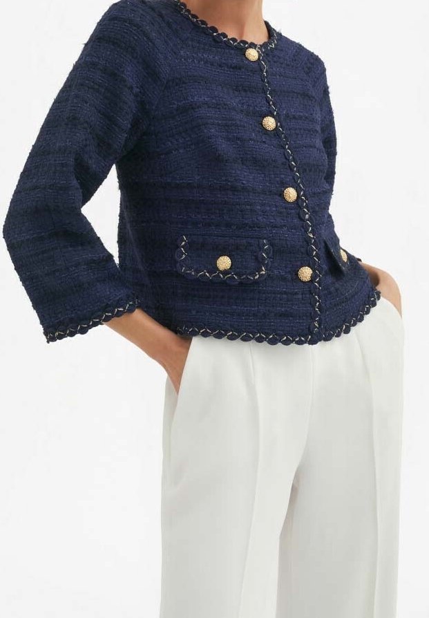 Kvinde iført en marineblå tekstureret jakke med guldknapper og hvide bukser, hænderne i lommerne, foran en hvid baggrund.