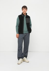 Fleece vest in het zwart met een rits, met een grijze borstzak. Gecombineerd met een groene longsleeve en een grijze broek, afgemaakt met beige schoenen.