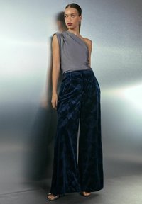 Woman leaning against a metallic wall wearing a one-shoulder gray top, dark blue patterned wide-leg pants, and black open-toe heels.