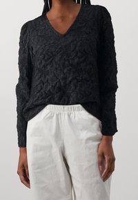 Blusa nera a trama con scollatura a V e maniche lunghe, indossata sopra pantaloni bianchi con cintura elastica. Leggero motivo a sgualcitura su tutta la superficie.