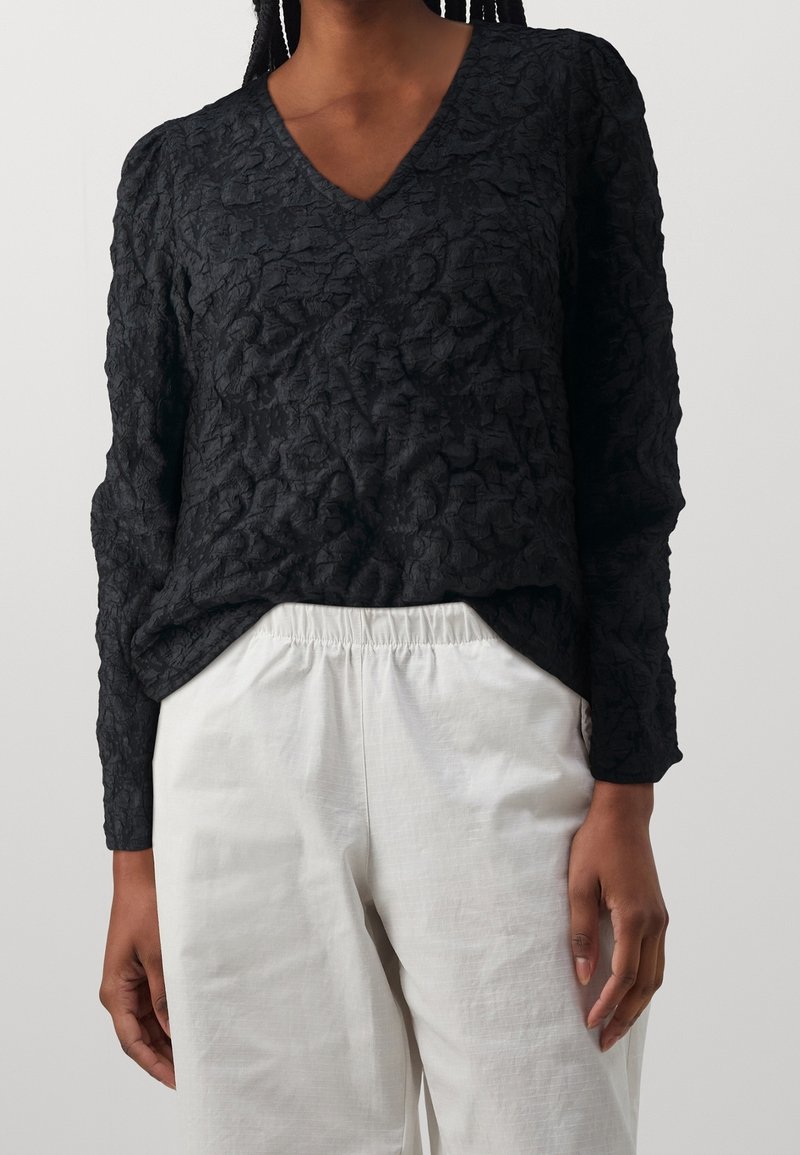 Blusa nera a trama con scollatura a V e maniche lunghe, indossata sopra pantaloni bianchi con cintura elastica. Leggero motivo a sgualcitura su tutta la superficie.