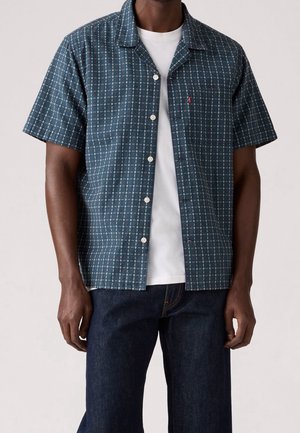 Homme portant une chemise à manches courtes ouverte, boutonnée, vert foncé à motifs, par-dessus un t-shirt blanc et un jean foné.