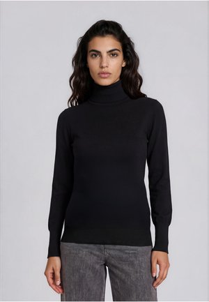 Vrouw met lang donker haar, die een zwarte coltrui en grijze spijkerbroek draagt tegen een effen lichtgrijze achtergrond.