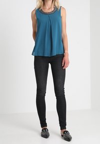 Turkos ärmlös blus med plisserad front och pärlbesatt krage, parat med svarta slim fit jeans och svarta platta skor med nitar.