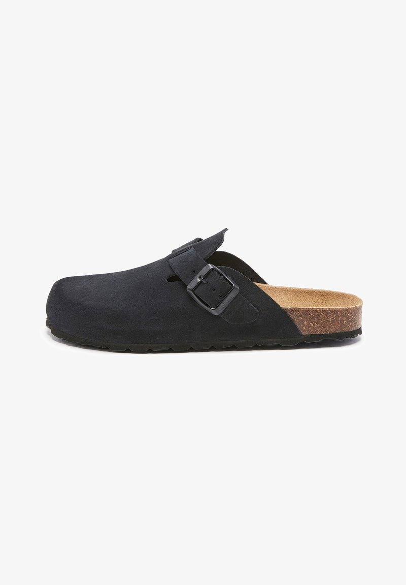 Sabot noir en suède à enfiler avec bride à boucle ajustable, semelle intérieure en liège et semelle extérieure en caoutchouc noir texturé, vu de côté sur fond blanc.