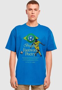 Blå bomulds-T-shirt med et design af det brasilianske flag i gult og grønt, trykt tekst og en illustration af en fodboldspiller.