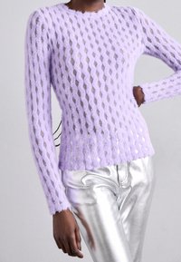 Kvinna som bär en långärmad lavendelfärgad fluffig stickad topp med ett diamanthål-mönster och blanka silvermetalliska byxor.