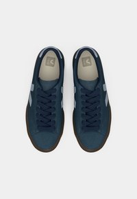 Tenisi bleumarin din suede cu sireturi de culoare albastru închis, accente albe ale logo-ului și talpă din cauciuc maro, având vârful rotunjit și guler căptușit.