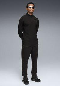 Chándal atlético negro con sudadera de cremallera de un cuarto, con un logo y bolsillos, combinado con zapatillas negras. El modelo lleva gafas de sol oscuras.