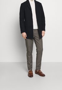 Schwarzer langer Mantel über einem hellen Pullover, kombiniert mit karierten grauen Hosen und braunen Loafers mit einer goldenen Schnalle als Akzent.