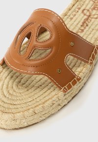 Smeđi kožni klizni sandal s izrezbarenim dizajnom, s jute užadom kao potplatom i detaljima šivanja. Okrogli oblik, teksturirana površina i naglasci od zlatne boje.