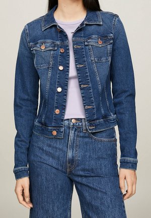 Vrouw die een donkerblauw spijkerjasje en bijpassende high-waisted jeans draagt, met een lichtpaarse top eronder, staande tegen een effen achtergrond.