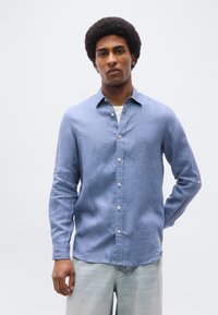 Νέος άνδρας με κοντό αφρο κούρεμα, ντυμένος με ανοιχτό μπλε πουκάμισο με κουμπιά και ανοιχτής απόχρωσης τζιν, στέκεται μπροστά σε απλό φόντο.