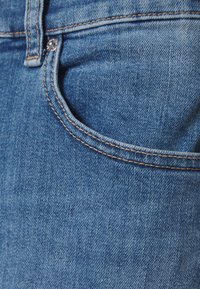 Denimtyg i ljusblått med en slät yta, med en framficka och kontrasterande bruna sömmar längs med kanterna.