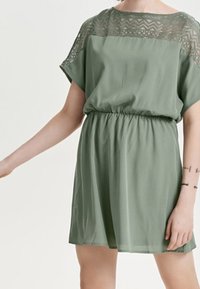 Vestido verde con mangas cortas, cintura elástica y detalles de encaje en el escote. Tejido suave y ligero con una silueta fluida.