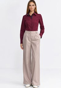 Bordowa koszula na guziki z kołnierzykiem, zestawiona z jasnobeżowymi spodniami w prążki. Model stoi z rękami w kieszeniach, ma na sobie białe buty.