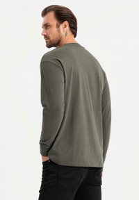 Mann mit mittellangen Haaren und einem Tattoo hinter dem Ohr, der ein olivgrünes Langarmhemd und schwarze Hosen trägt und seitlich schaut.